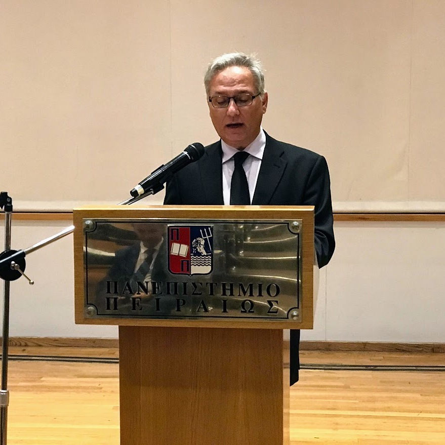 Author profile pic of Constantinos Koliopoulos in a dark-coloured suit, standing at a podium as if giving a speech